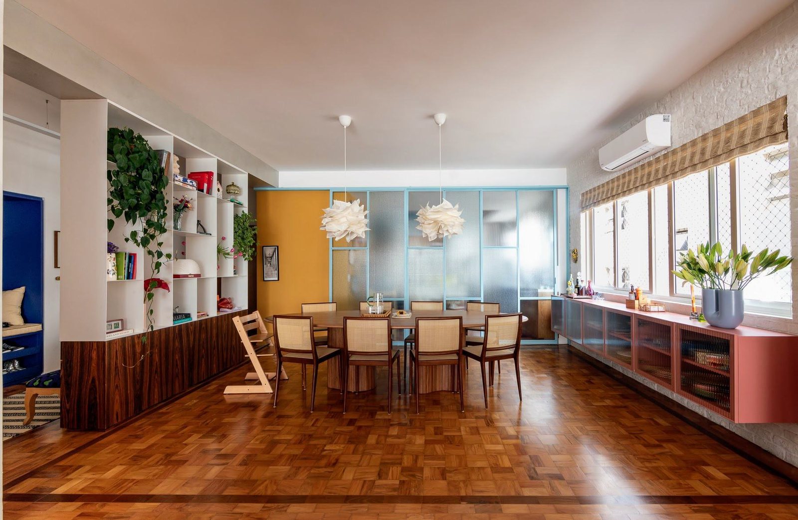 Sala de jantar com mesa e cadeiras de palhinha, parede amarela, estante branca e piso de tacos de madeira