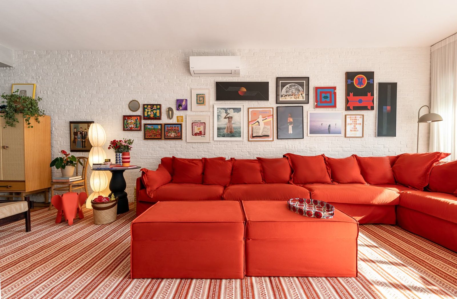 Sala de estar com sofá vermelho em L, parede de tijolos brancos coberta de quadros e tapete listrado
