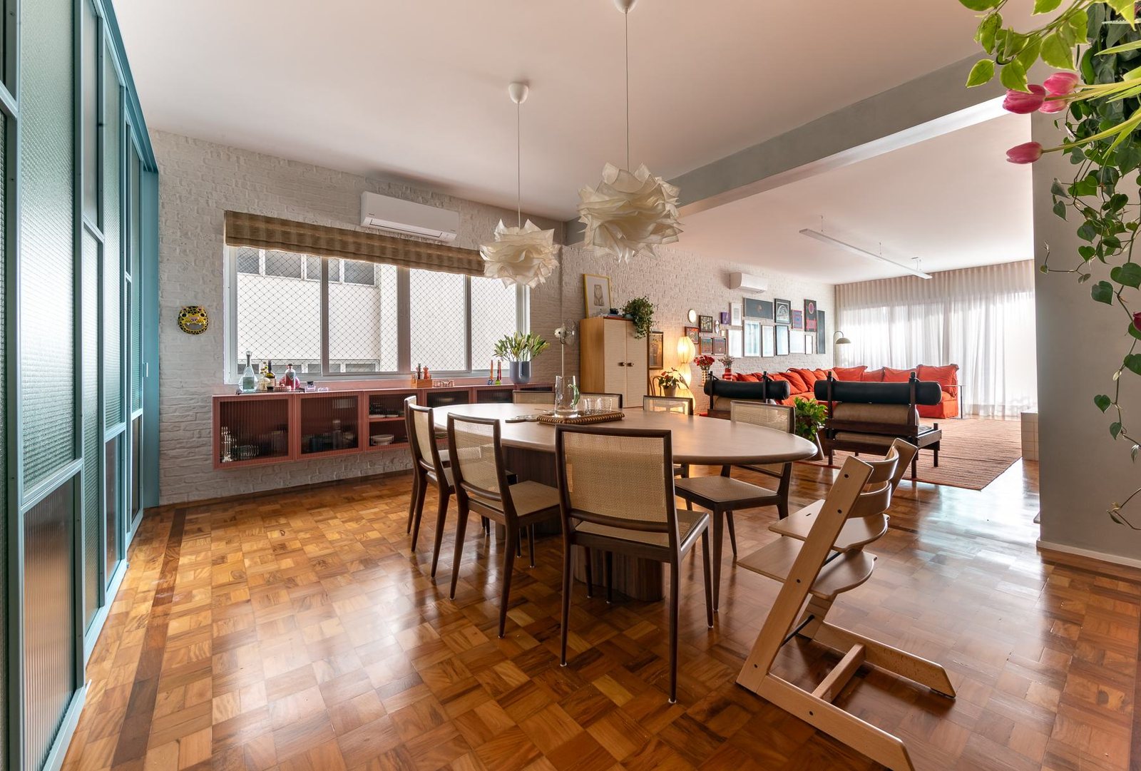 Sala de jantar com mesa redonda, cadeiras de madeira, plantas pendentes e sala de estar ao fundo