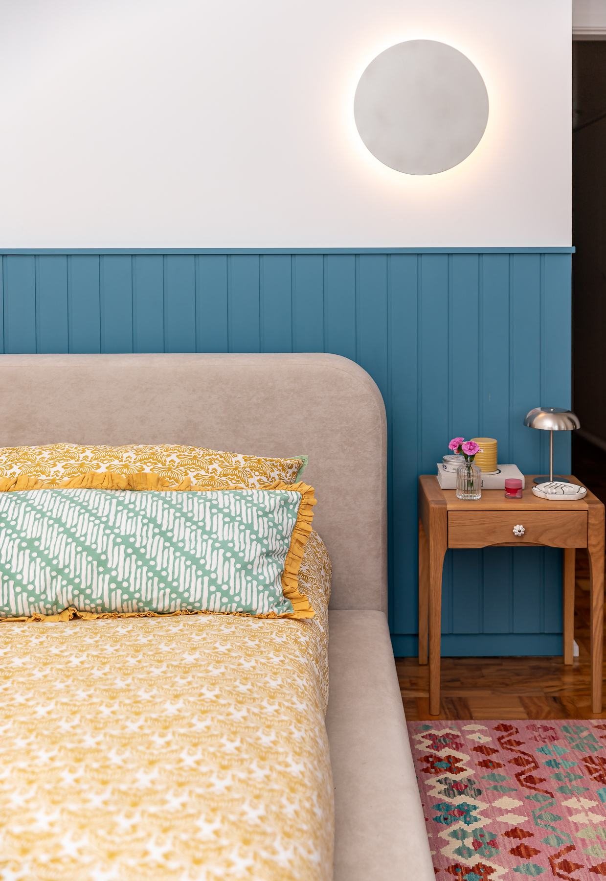 Quarto com cabeceira e lambri azul, roupa de cama amarela e criado-mudo de madeira