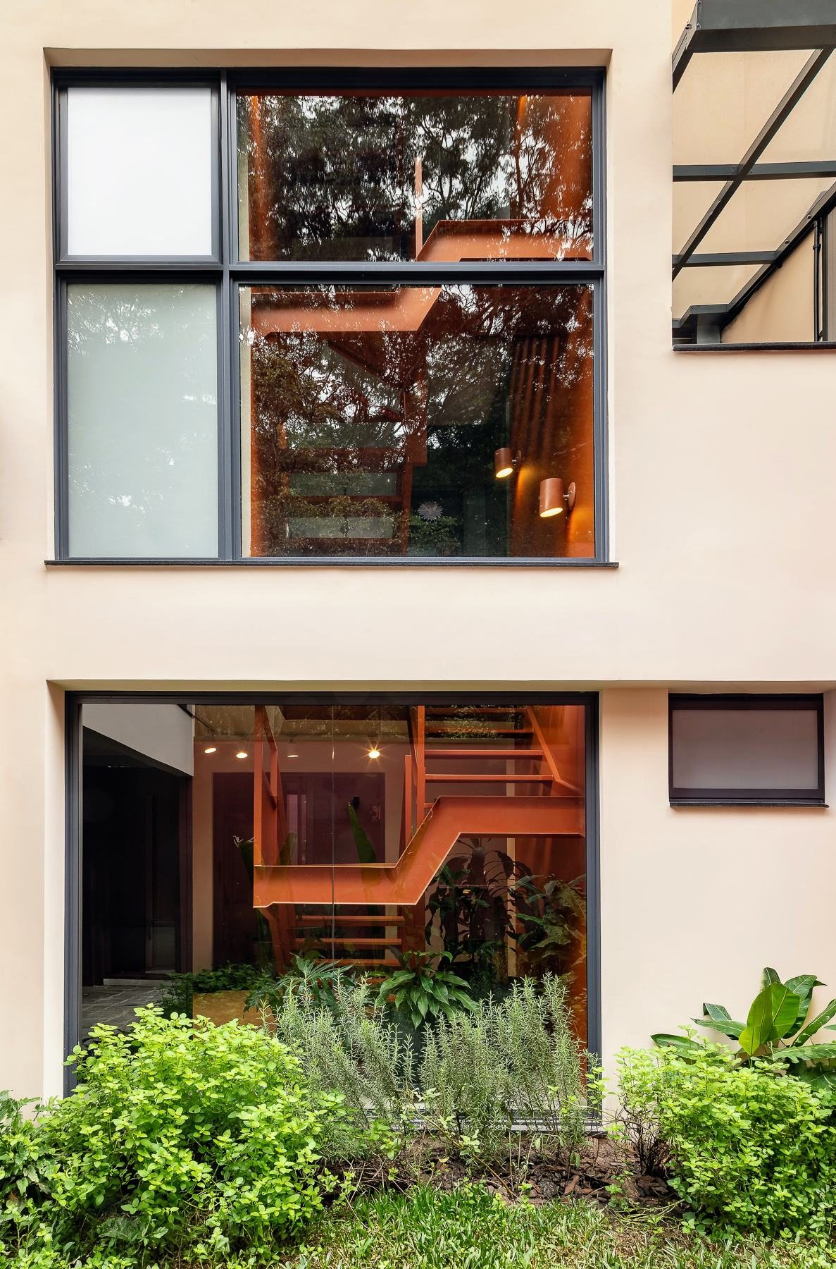 Fachada de casa com janela de esquadria escura refletindo árvores e escada laranja vista através do vidro, com jardim ao pé