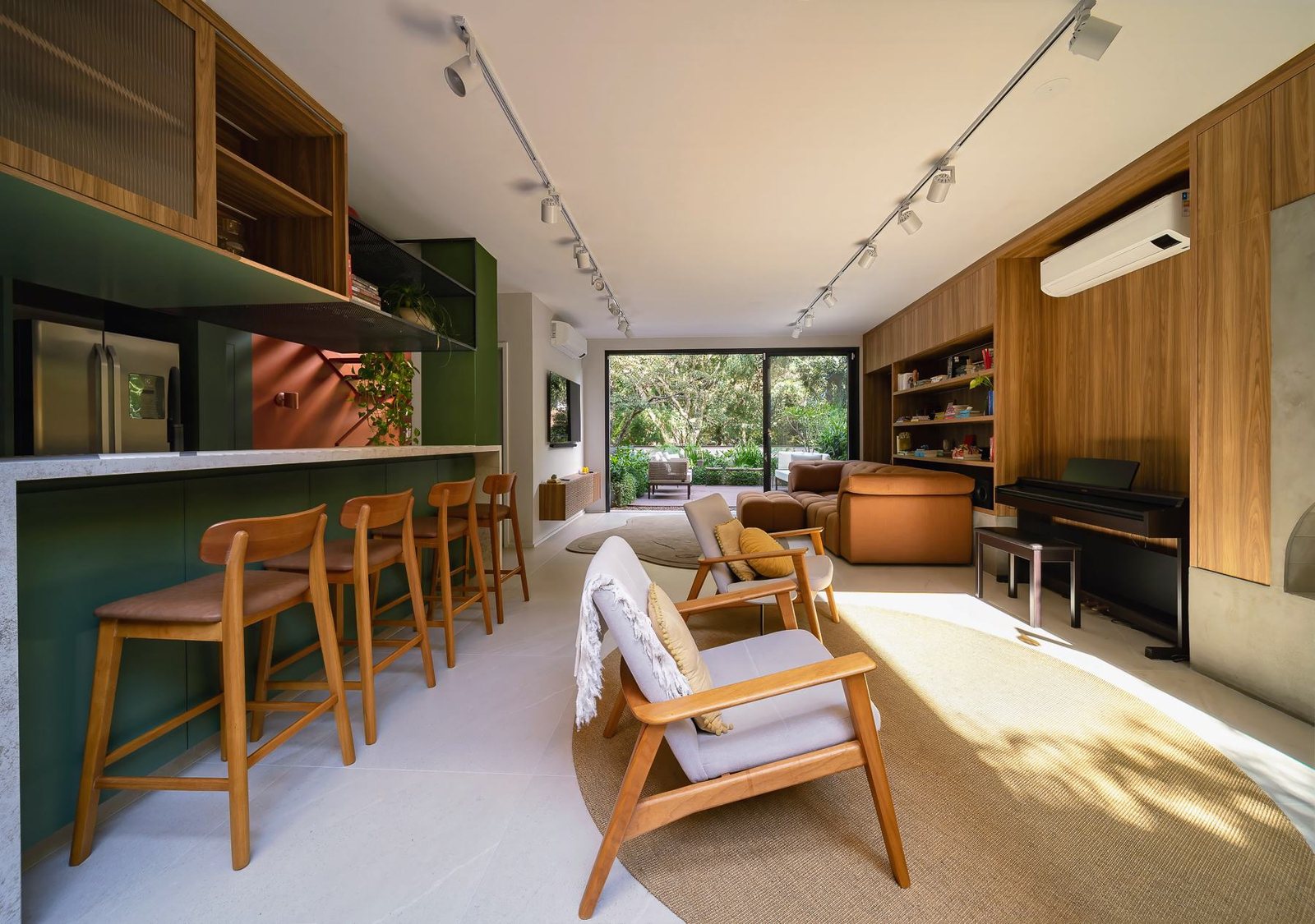 Ambiente integrado com bancada e banquetas de madeira em frente à cozinha verde e sala de estar com poltronas, tapete e piano ao fundo