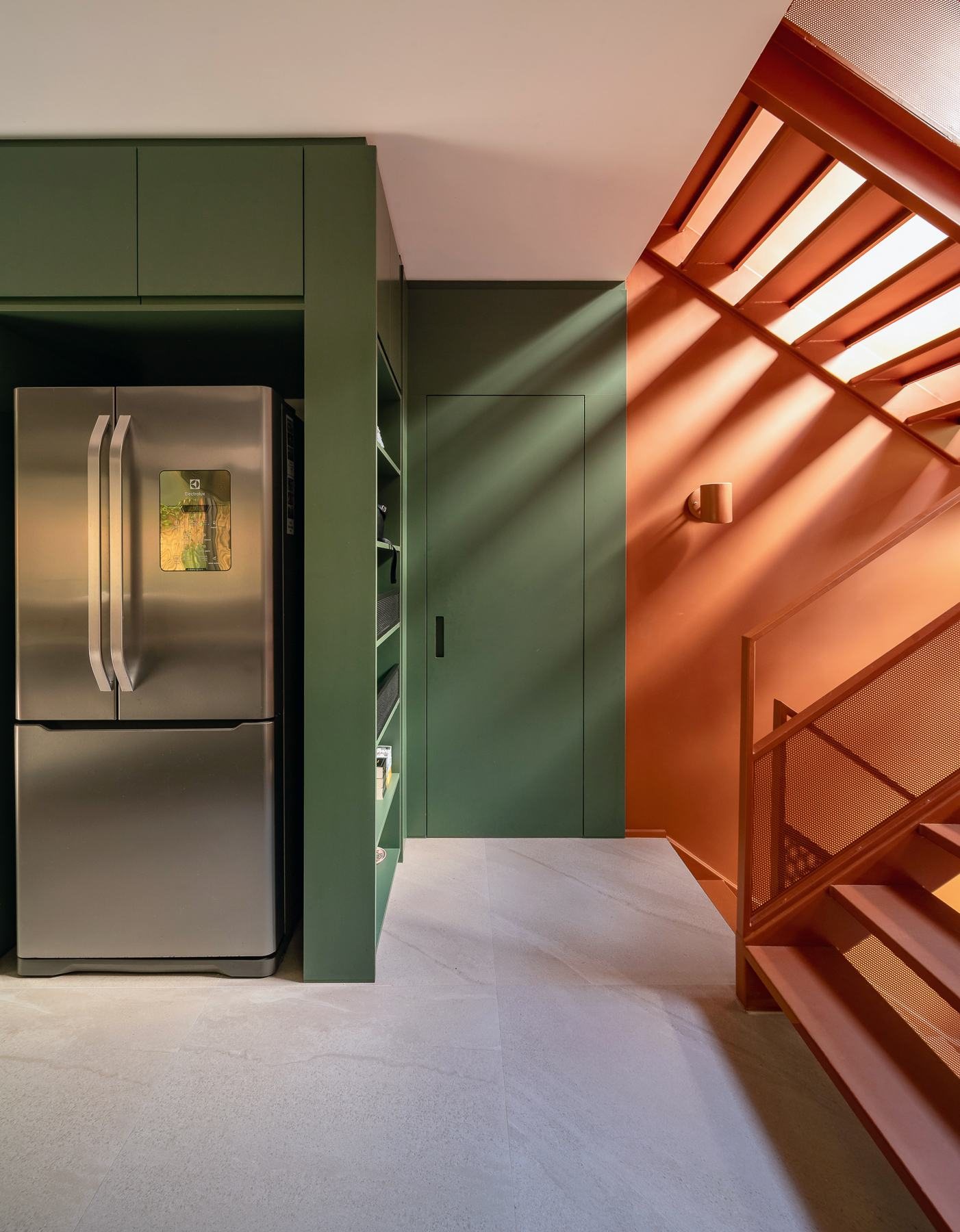 Geladeira inox junto à parede verde, com a escada laranja ao fundo