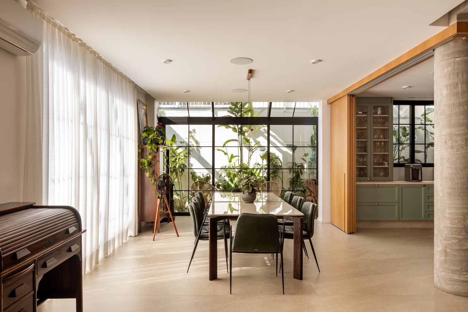 Sala de jantar com mesa e cadeiras verdes, cortinas claras, plantas e coluna de concreto, com cozinha verde ao fundo