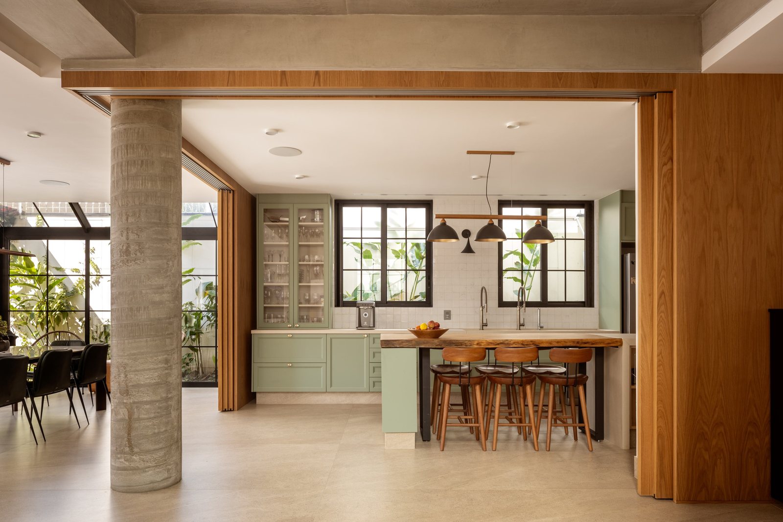 Cozinha ampla com coluna de concreto redonda, marcenaria verde-provençal, ilha de madeira com banquetas e grandes esquadrias pretas