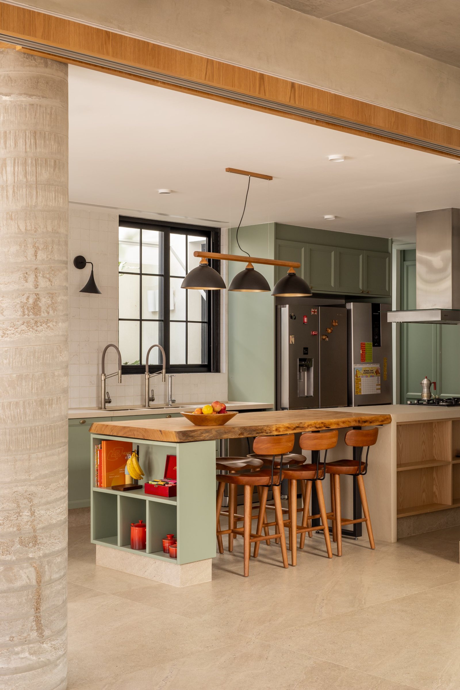 Cozinha verde com ilha, banquetas, pendentes e geladeira ao fundo, junto à coluna de concreto
