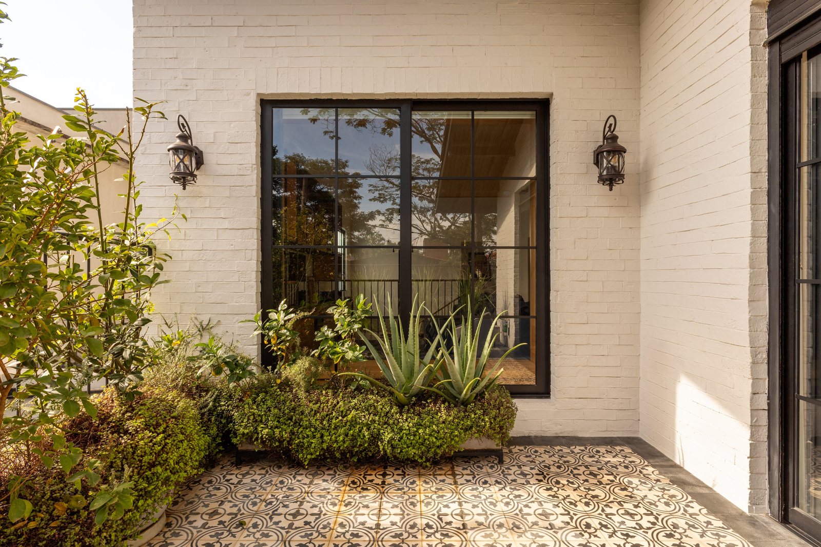 Pátio de casa com parede de tijolo branco, grande porta de vidro com esquadria preta, agave e canteiro de plantas sobre piso de ladrilho hidráulico